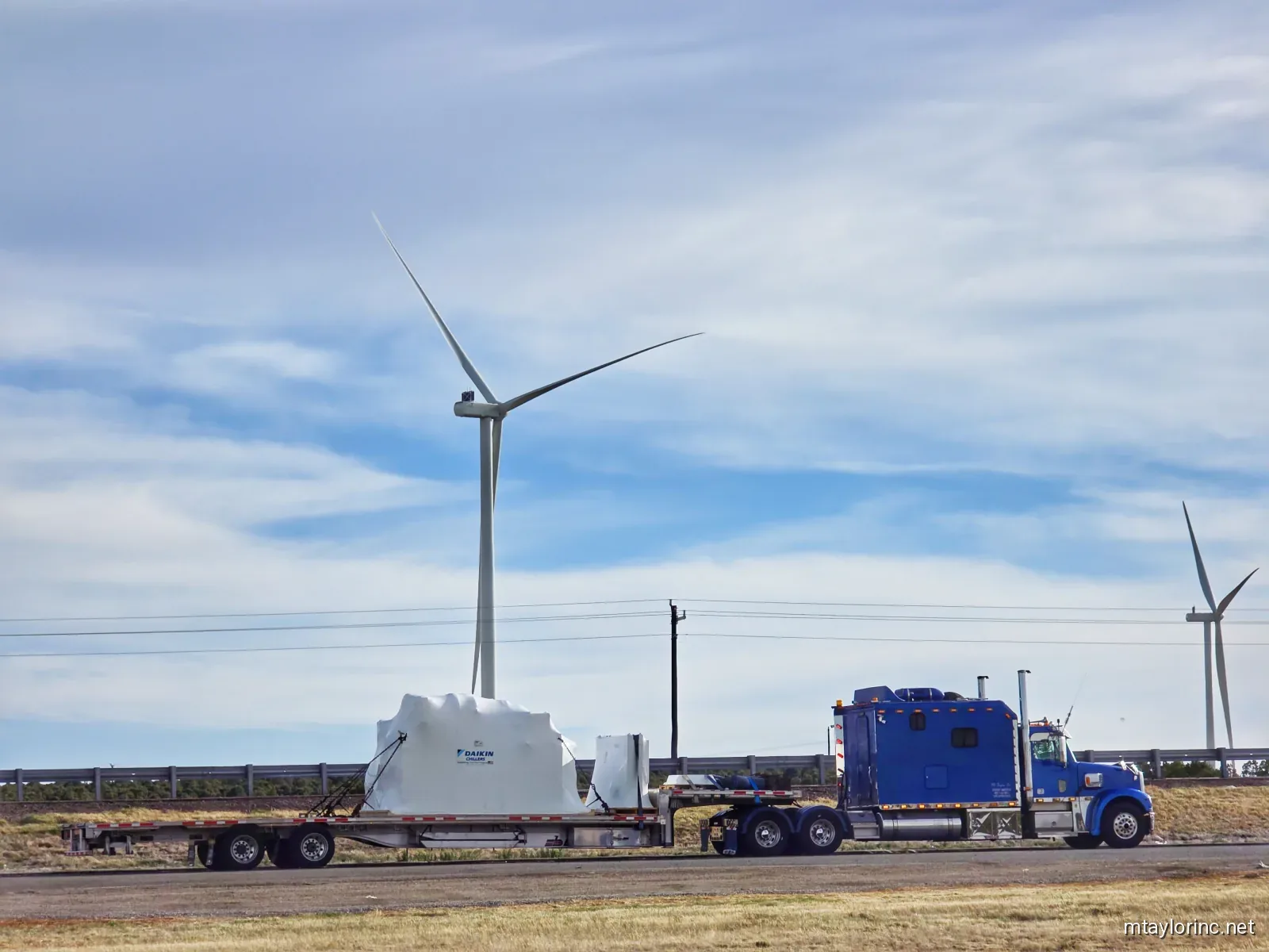 M Taylor Inc stepdeck hauling Daikin HVAC equipment past wind turbines